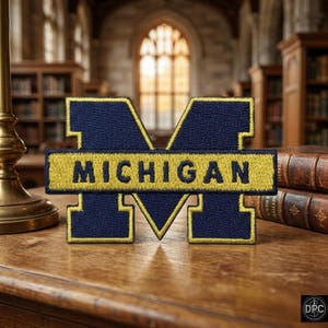 Peut inclure: Un écusson brodé bleu marine et jaune représentant une grande lettre M en bloc avec le mot MICHIGAN cousu en bleu marine sur une barre horizontale jaune qui recouvre le centre de la lettre M, fini avec une bordure jaune.