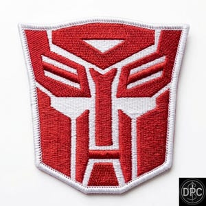 May include: A shield-shaped embroidered patch featuring the red and white Autobot insignia from the Transformers franchise. The design consists of a stylized robotic face with geometric red segments outlined and separated by white thread.