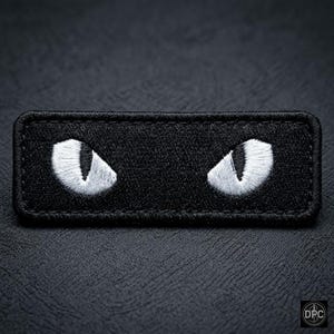 May include: A rectangular black patch embroidered with two white cat eyes. The eyes are stylized with a sharp, angular design. The patch has a stitched border and is set against a textured black background.