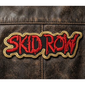 May include: Embroidered patch featuring the text "SKID ROW" in red, outlined in gold, on a black background. The patch is sewn onto a brown leather jacket, showcasing a vintage aesthetic.