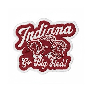 May include: A red and white embroidered patch featuring the word "Indiana" in cursive above a stylized bison illustration. Below the bison, the text "Go Big Red!" is displayed in white cursive lettering. The patch has a white border.