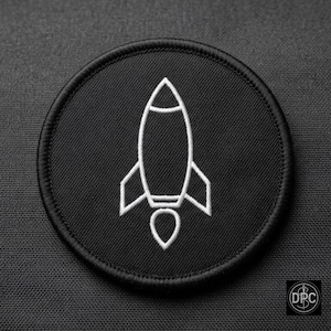May include: A circular black embroidered patch featuring a white line art design of a rocket ship. The rocket is centered on the patch, which has a thick black stitched border and a textured fabric surface throughout the entire circular shape.