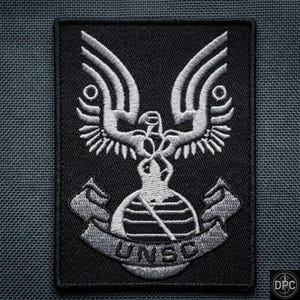 May include: A rectangular black embroidered patch featuring a silver-colored emblem of an eagle with spread wings perched atop a globe. Below the globe, a curved banner displays the text UNSC in bold, capitalized silver-colored lettering.