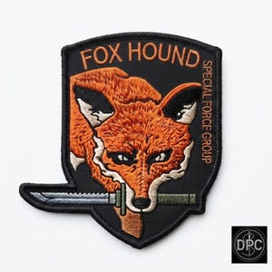 May include: A shield-shaped embroidered patch with a black background featuring an orange fox head holding a serrated knife in its mouth. The text FOX HOUND is arched at the top, and SPECIAL FORCE GROUP is written vertically along the right side.