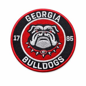 May include: Embroidered patch featuring the Georgia Bulldogs logo. The patch is circular, with a black outer ring and a red inner circle. The logo includes a bulldog head, the words "GEORGIA BULLDOGS," and the numbers "17" and "85."