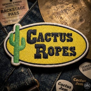 May include: An oval-shaped embroidered patch with a yellow background and a white border. The patch features a green cactus on the left side and the black text CACTUS ROPES in the center. The embroidery has a textured finish throughout.
