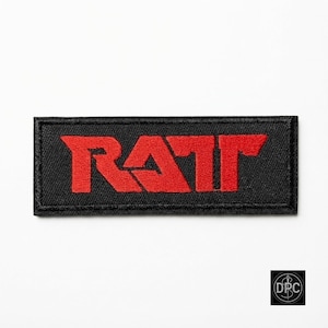May include: A rectangular embroidered patch featuring a black fabric background with the word RATT stitched in bold red thread. The lettering has a distinct, sharp-edged font style, and the entire patch is bordered by a thick black stitched edge.