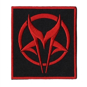 May include: Square black patch with a red embroidered design. The design features a stylized red emblem within a red circle, with sharp, angular shapes. The patch has a red border.