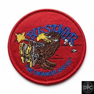 May include: A circular embroidered patch with a red background featuring a brown creature with a yellow beak and yellow boots. The text BUG STOMPER is in blue above the creature, and the text WE ENDANGER SPECIES is in blue below it.