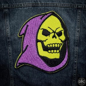 May include: An embroidered patch featuring a yellow skull with black eye sockets and teeth, wearing a purple hood with black outlines. The patch is stitched onto a dark blue denim fabric background. The design depicts a skeletal character head.