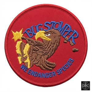 May include: A circular embroidered patch with a red background. It features a brown eagle with a yellow beak and talons, a yellow starburst, and blue text that reads BUG STOMPER and WE ENDANGER SPECIES. The patch has a red stitched border.