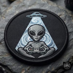 Op de afbeelding: Een ronde geborduurde patch met een grijze alien die in kleermakerszit zit en een videogamecontroller vasthoudt. Boven de alien zendt een UFO een lichtstraal uit met een wervelend blauw en wit patroon tegen een zwarte achtergrond.