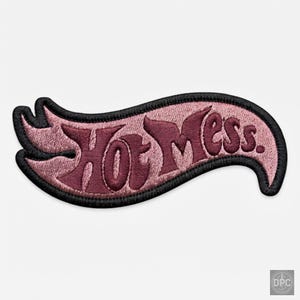 Può includere: Una toppa ricamata con sfondo rosa chiaro e bordo nero, a forma di fiamma stilizzata. Il testo Hot Mess. è cucito con filo bordeaux scuro al centro della toppa in uno stile di carattere audace e fluido.