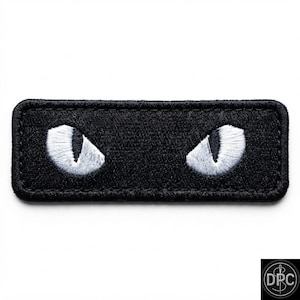 May include: A rectangular embroidered patch featuring a solid black background with two stylized white cat eyes. The eyes are almond-shaped with vertical black pupils, centered horizontally within the black fabric border of the patch.