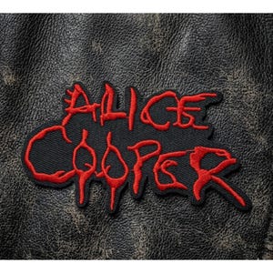May include: Black embroidered patch with the name "ALICE COOPER" in red, stylized lettering. The patch is rectangular with a black background and is set against a textured, dark leather surface.