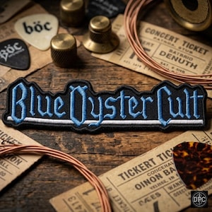 May include: A rectangular embroidered patch featuring the text Blue Oyster Cult in light blue gothic lettering on a black background, with a thin white horizontal line running underneath the text, designed for attachment to clothing or fabric items.
