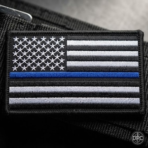 May include: A rectangular embroidered patch featuring a stylized American flag design in black, white, and grey tones. The flag includes a single horizontal blue stripe running across the center, with white embroidered stars on a black field.