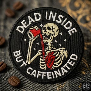 May include: A circular black embroidered patch featuring a white skeleton drinking from a red cup. The liquid flows from the cup into a red anatomical heart in the chest cavity. The text reads DEAD INSIDE BUT CAFFEINATED in white letters.