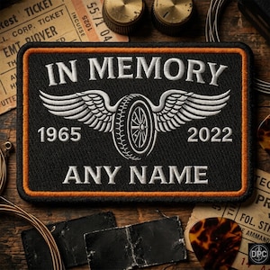 May include: A rectangular black embroidered patch with an orange border. It features the white text IN MEMORY, a central winged tire graphic, the years 1965 and 2022, and the text ANY NAME in white embroidery on a black fabric background.