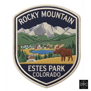 May include: A shield-shaped embroidered patch featuring a landscape of Rocky Mountain National Park. The design includes snow-capped mountains, a blue lake, green pine trees, and a brown moose. Text reads ROCKY MOUNTAIN ESTES PARK COLORADO.