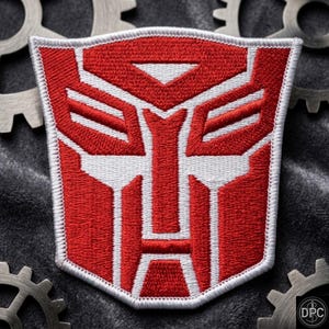 May include: A shield-shaped embroidered patch featuring the red and white Autobot insignia from the Transformers franchise. The design consists of a stylized robotic face with geometric lines and a thick white border, showcasing detailed thread work.
