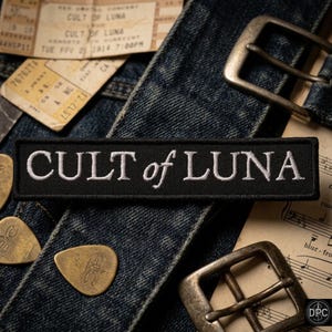 May include: A rectangular black embroidered patch featuring the white text CULT of LUNA in a serif font. The patch is positioned over a dark blue denim fabric with metal belt buckles, guitar picks, and vintage paper tickets visible nearby.