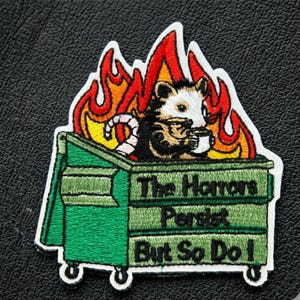 Op de afbeelding: Geborduurde patch met een opossum in een brandende vuilnisbak, met een mok. De patch heeft de tekst "The Horrors Persist But So Do I" in groen en wit. De patch staat op een zwarte achtergrond.