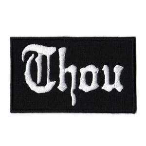 May include: A rectangular black embroidered patch featuring the word Thou in white gothic-style lettering. The patch has a clean, stitched border and a flat, uniform surface, showcasing high-contrast white thread against a solid black fabric background.