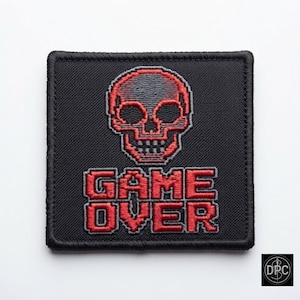 May include: A square black embroidered patch featuring a red pixelated skull graphic positioned above the red pixelated text GAME OVER. The patch has a thick black stitched border and a textured fabric surface with detailed red and grey thread work.