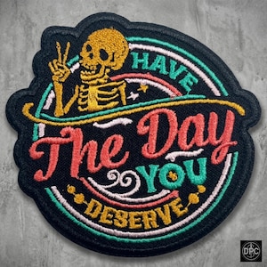 Könnte beinhalten: Gestickter Aufnäher mit schwarzem Hintergrund, der ein gelbes Skelett zeigt, das ein Friedenszeichen gibt. Der Aufnäher trägt die Worte "Have The Day You Deserve" in einer farbenfrohen, stilisierten Schriftart, mit einem Regenbogen und Sternen.