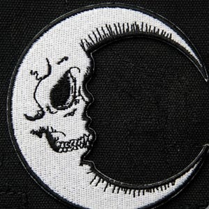 May include: Embroidered patch featuring a white crescent moon with a skull design on a black background. The skull is detailed with eye sockets, a nose, and teeth. The moon's edges are outlined with a white border.