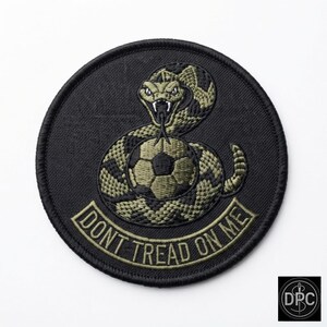 Può includere: Una toppa ricamata circolare con sfondo nero raffigurante un serpente arrotolato di colore verde oliva e nero attorno a un pallone da calcio. Sotto il serpente, un nastro verde oliva contiene il testo DONT TREAD ON ME in lettere nere.