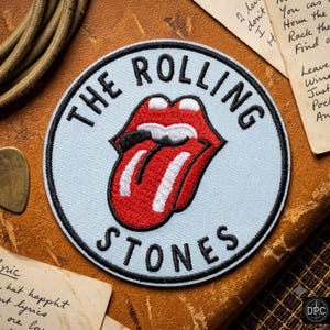 May include: A circular embroidered patch featuring a light blue background with a black border. The iconic red lips and tongue logo is centered, with the black text THE ROLLING STONES arched around the top and bottom of the design.