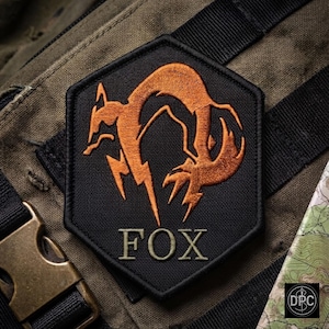 Könnte beinhalten: Ein sechseckiger, schwarzer Stickaufnäher mit einem stilisierten orangefarbenen Fuchsemblem und dem Wort FOX, das in hellgrünem Garn unter der Grafik gestickt ist. Der Aufnäher hat einen dicken schwarzen Rand und ist auf olivgrünem Stoff.