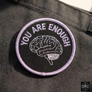 Op de afbeelding: Een ronde geborduurde patch met een zwarte achtergrond en een lichtpaarse rand. De patch bevat een witte lijntekening van een menselijk brein in het midden, met de tekst "YOU ARE ENOUGH" geborduurd in lichtpaarse letters.