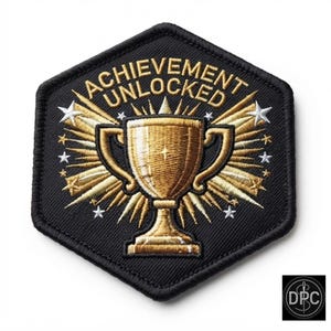 May include: A hexagonal embroidered patch with a black background and a gold trophy in the center. The text "ACHIEVEMENT UNLOCKED" is arched in gold thread above the trophy. Gold rays and small white stars radiate from behind the trophy.