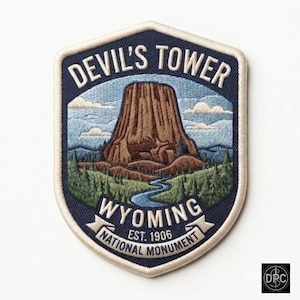 May include: A shield-shaped embroidered patch featuring a brown Devil's Tower monument in Wyoming, surrounded by green pine trees and a blue river. The text reads DEVIL'S TOWER, WYOMING, EST. 1906, and NATIONAL MONUMENT in white lettering.