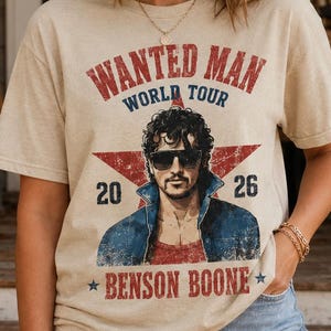 May include: A beige short-sleeve t-shirt featuring a graphic print of a man in sunglasses and a denim jacket. The text reads WANTED MAN WORLD TOUR 2026 and BENSON BOONE, centered over a large red star. The design has a distressed, vintage aesthetic.