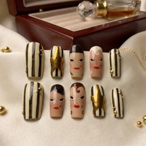 May include: A set of ten press-on nails with various designs. Some nails feature vertical black and white stripes, others have gold star accents, and some are painted with faces. Gold beads and a gold chain with a star charm are also present.