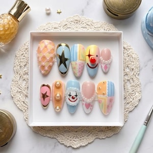 May include: A set of ten colorful, decorated false nails in a white tray. Designs include stars, clown faces, and geometric patterns. The nails are adorned with pearls and other embellishments. A bottle of perfume and other beauty accessories are in the background.