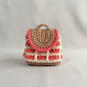 May include: A small, handcrafted crochet backpack featuring a textured pattern in shades of brown, cream, and muted red. The bag has a rounded top flap with a matching handle, a structured base, and a grid-like stitch design across the front.