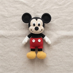 May include: A handmade crochet doll of a mouse character with large round black ears, a cream-colored face, and a black nose. The doll wears red shorts with two white buttons, white gloves, and yellow shoes, all crafted with a textured yarn stitch.