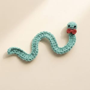 May include: A light green crocheted worm toy featuring a wavy, segmented body, two small black bead eyes, and a small red crocheted bow tie attached just below the head. The yarn texture is visible throughout the entire length of the object.