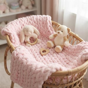 May include: A light pink chunky knit blanket draped inside a round wicker bassinet. A plush cream-colored bunny toy holding a wooden ring and a small beige teddy bear sit on the blanket alongside a smooth wooden heart-shaped object.