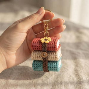 May include: A crochet keychain featuring a stack of three miniature books in terracotta, beige, and teal. The books are bound together by a dark brown crocheted strap with a small wooden button and a yellow crocheted flower on top.