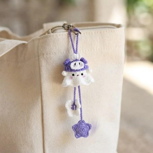 Puede incluir: Un llavero de fantasma tejido a ganchillo con cuerpo blanco y gorro morado con hocico de cerdo. El dije cuelga de un cierre de metal plateado e incluye dos accesorios tejidos colgantes: una pequeña flor blanca y una estrella morada de cinco puntas.