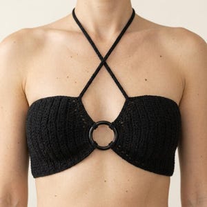 Puede incluir: Un top halter de ganchillo negro con un patrón de punto texturizado y un detalle de anillo circular central. La prenda tiene tirantes finos y cruzados que se extienden desde el anillo central hasta el cuello, creando una silueta estructurada.