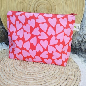 May include: A red cosmetic bag patterned with pink hearts. It features a red zip and a small tag with text. The bag rests on a woven mat.