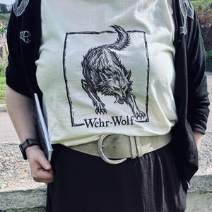 May include: A cream-coloured t-shirt featuring a black ink-style illustration of a snarling wolf inside a rectangular frame, with the text "Wehr-Wolf" printed below the graphic. The shirt is worn with a black cardigan, a wide beige belt, and a black skirt.