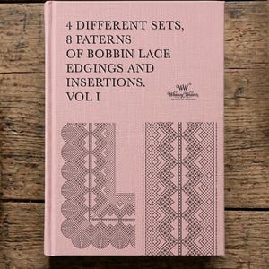 May include: A pink book titled "4 Different Sets, 8 Patterns of Bobbin Lace Edgings and Insertions. Vol I." The cover features black lace patterns and the "Whimsy Weaver" logo. The book is on a wooden surface.
